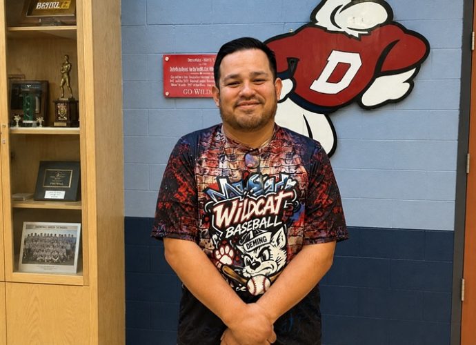 Deming Public Schools Athletic Director Nelson Diaz stands in front of a Wildcat logo inside the Deming High School athletics office.