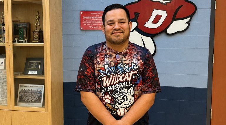 Deming Public Schools Athletic Director Nelson Diaz stands in front of a Wildcat logo inside the Deming High School athletics office.