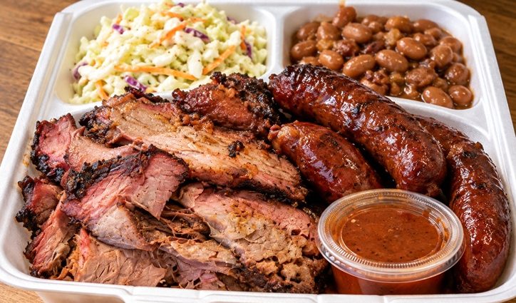 A takeout meal of klobase, barbecue meat, coleslaw and pinto beans with a cup of BBQ sauce.
